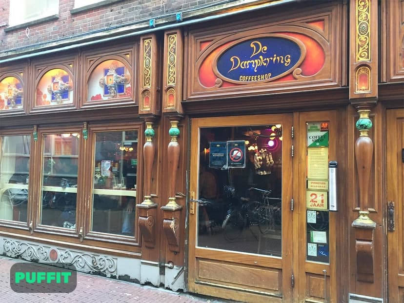 שלט הזכוכית הצבעוני של De Dampkring Coffeeshop מעל חלונות העץ המגולפים ברחוב